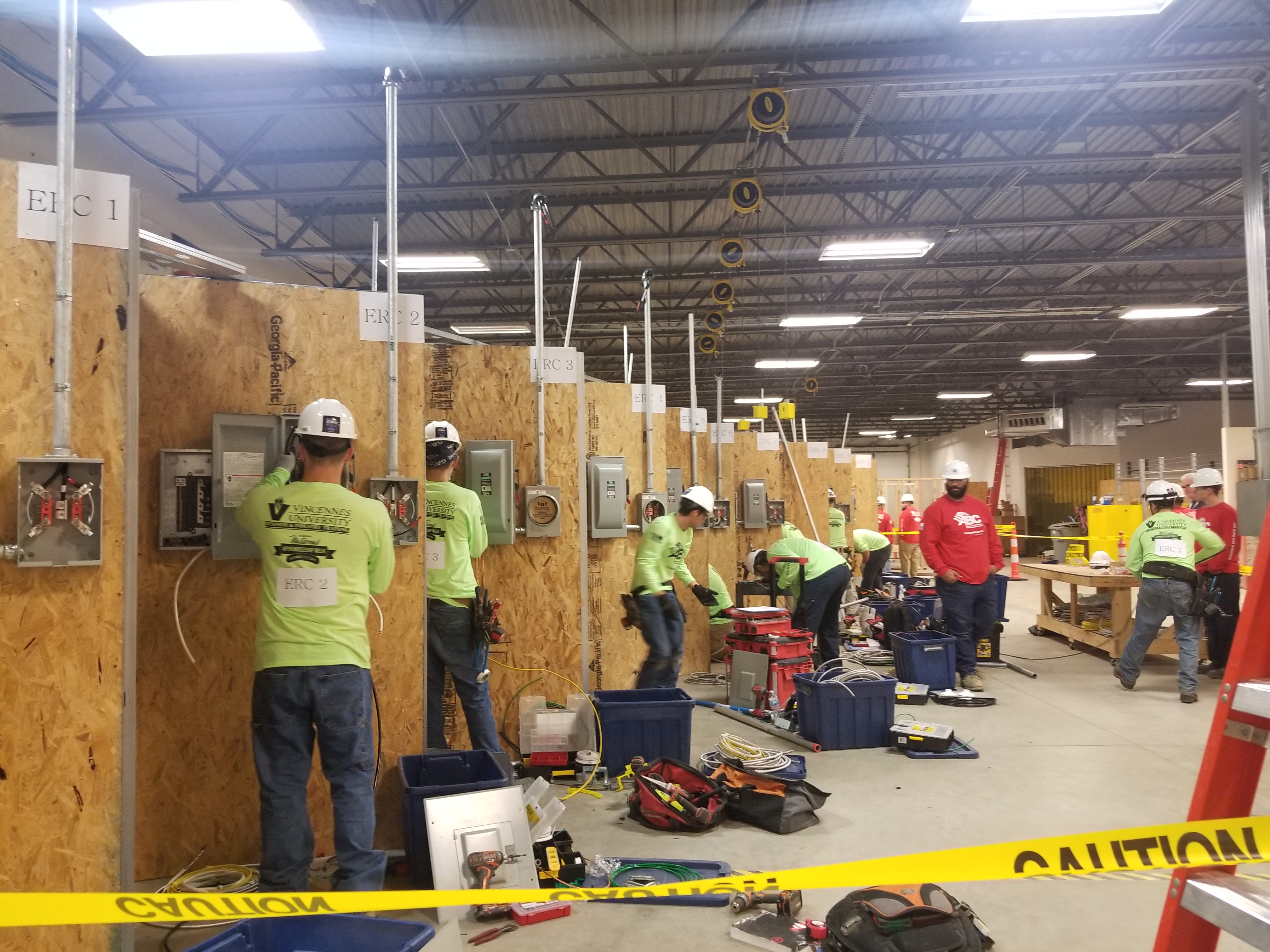 People in hard hats work at individual electrical panels with equipment around them while Instructors look on.