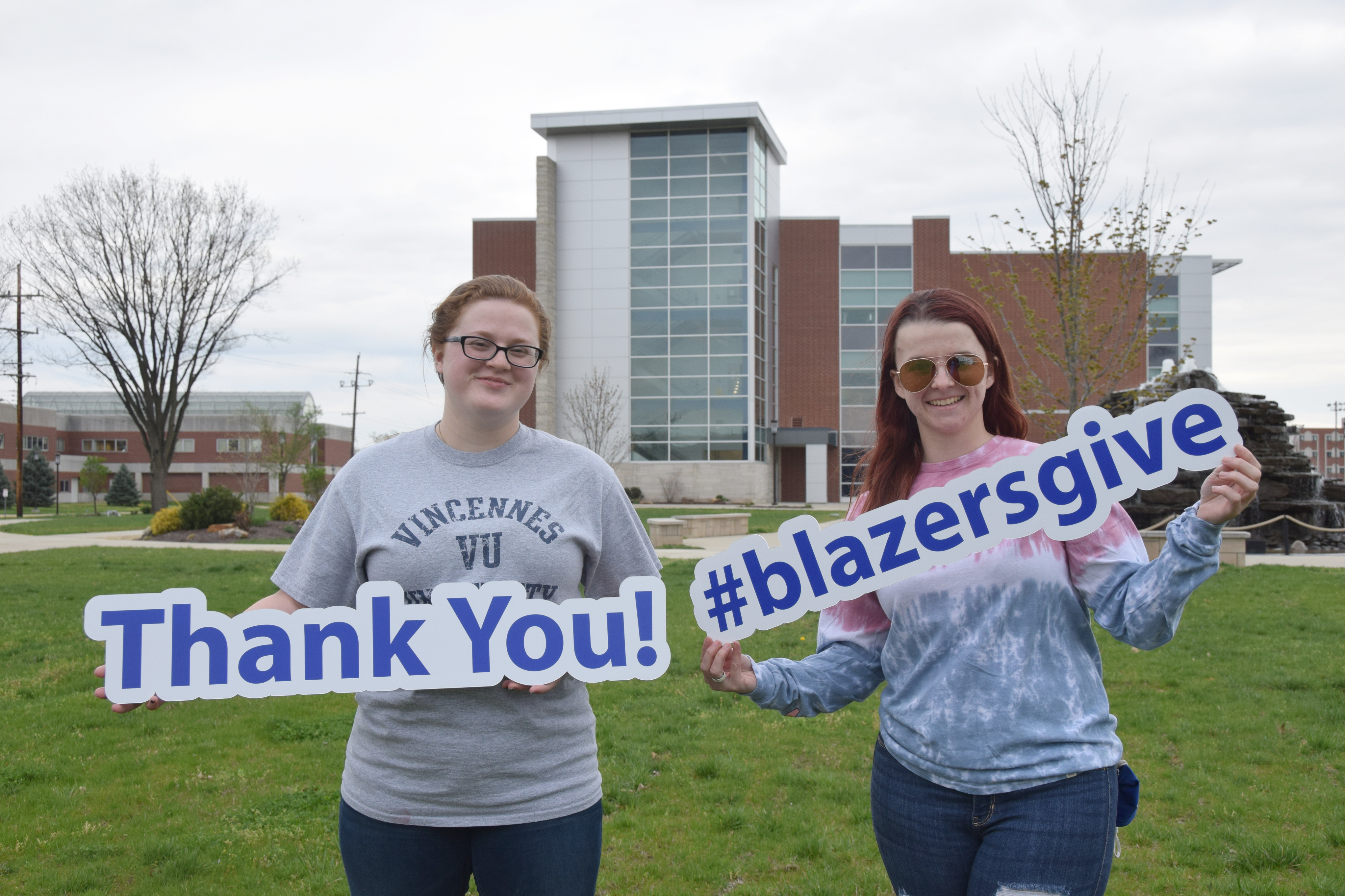 Two female VU students holding signs that say thank you and #blazersgive in front of fountain and Updike Hall on Vincennes Campus.